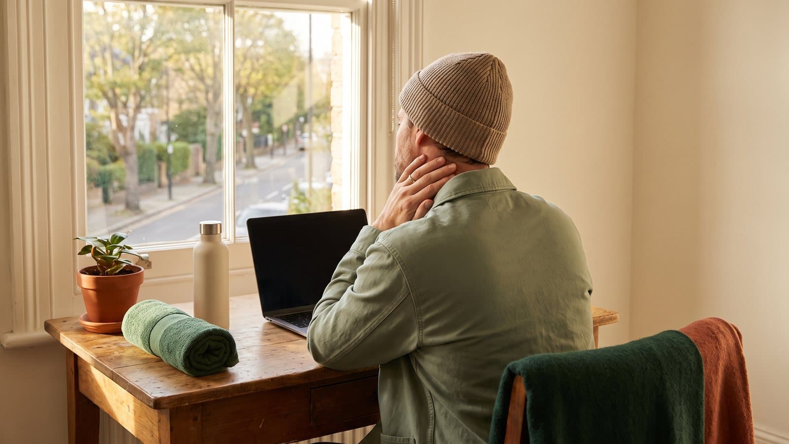 A person seen from behind at a sunlit desk pauses mid-task, one hand resting along the jaw, a water bottle beside the laptop