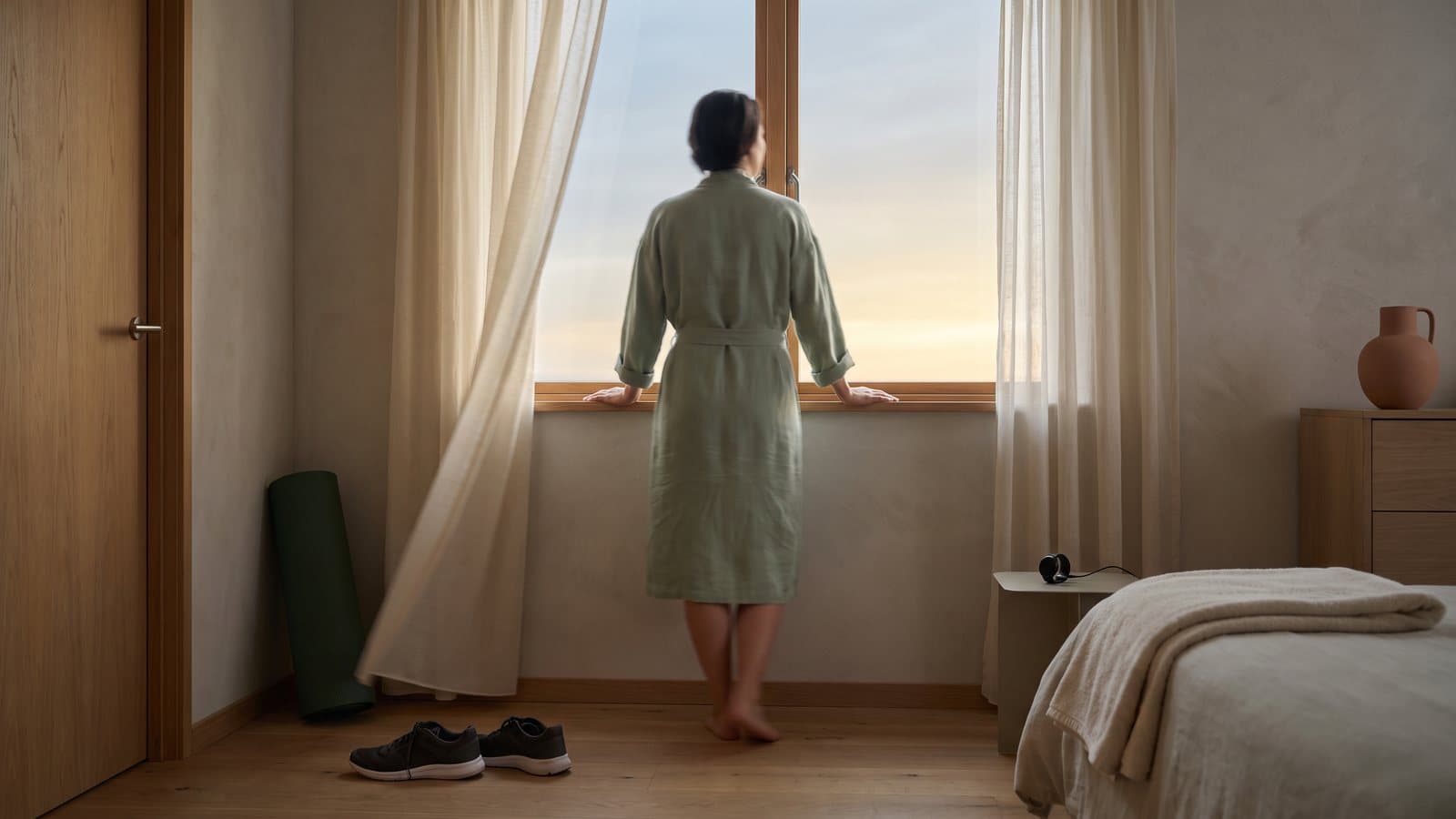 An adult stands at an open window at dawn taking a slow breath, running shoes waiting by the door