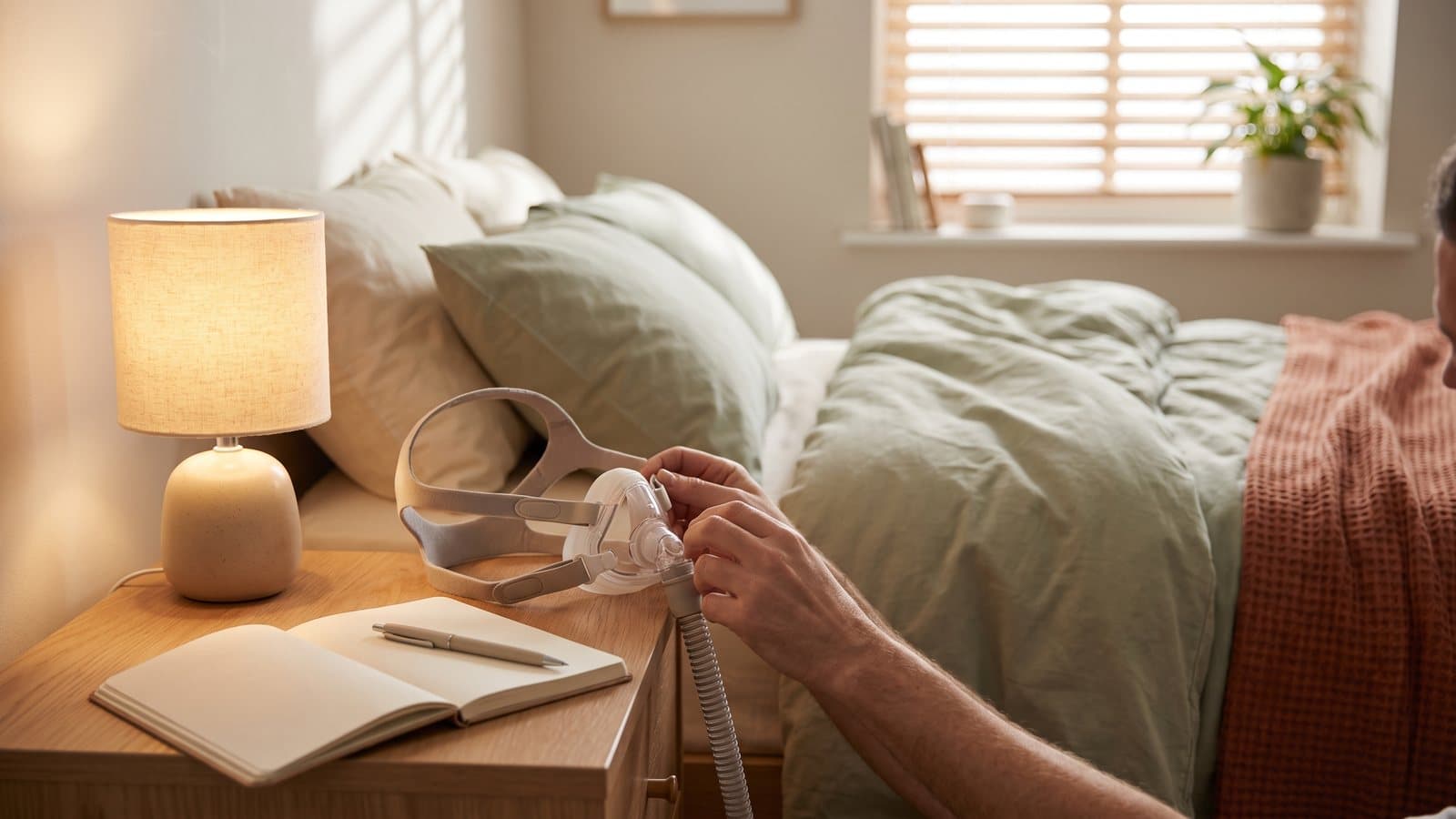 Hands adjust a CPAP mask strap on a nightstand beside a notebook, lit by a small lamp in early morning light